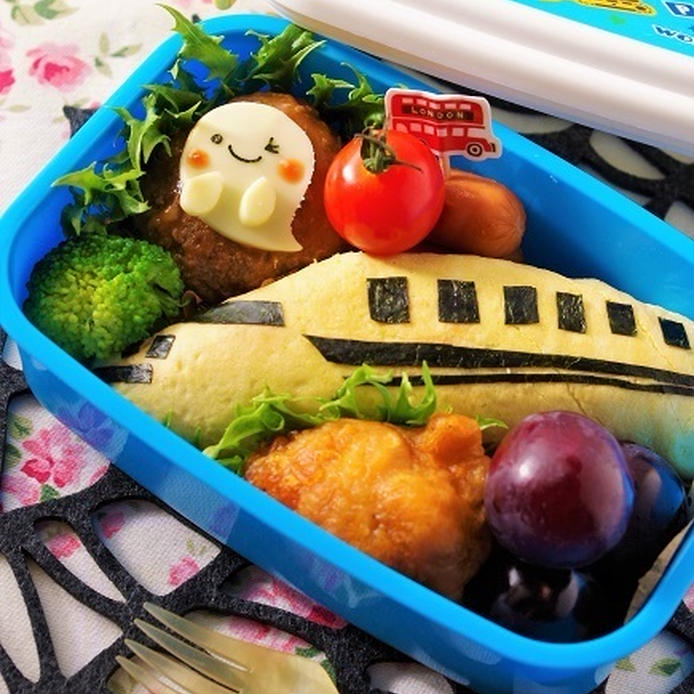 青いお弁当箱の中に、薄焼き卵で包まれた新幹線型のご飯が置かれ、そのまわりには唐揚げとプチトマトと巨峰が詰められている。