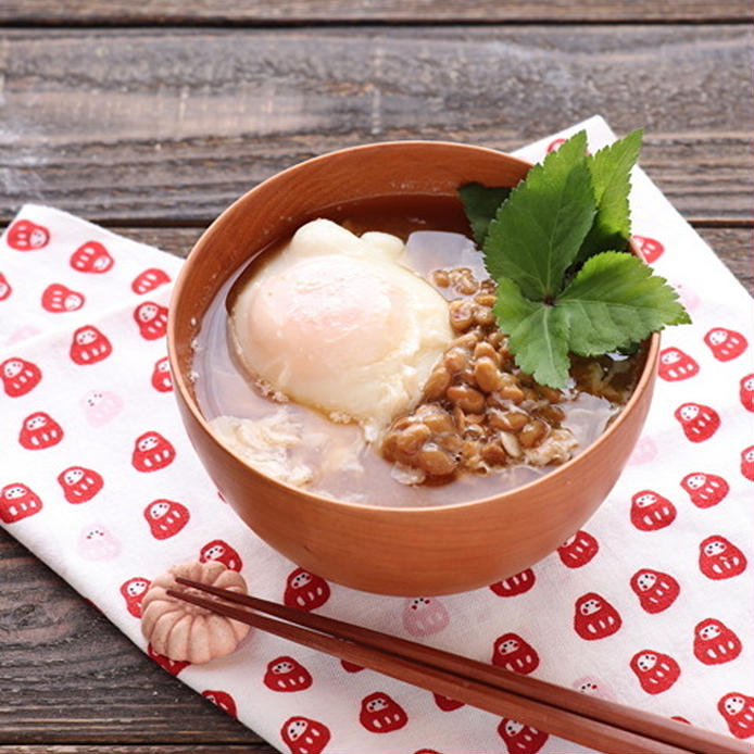 だるま柄のナプキンの上にある茶色の汁椀に盛られている納豆と落とし卵の味噌汁