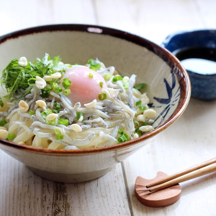 丼に入った温玉のせしらすうどんに、猫型箸置きに置かれた箸と、青い小鉢に入ったしょうゆが添えられている